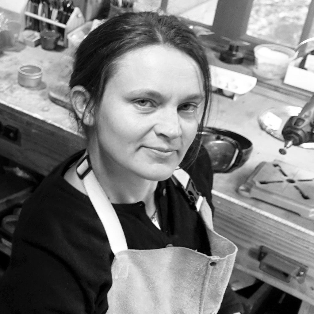 Artist LothLorien Stewart in her jewelry studio, wearing a metalsmith's apron at her workbench surrounded by jewelry-making tools and materials. Black and white portrait of the Mostly Sweet Jewelry creator at work.