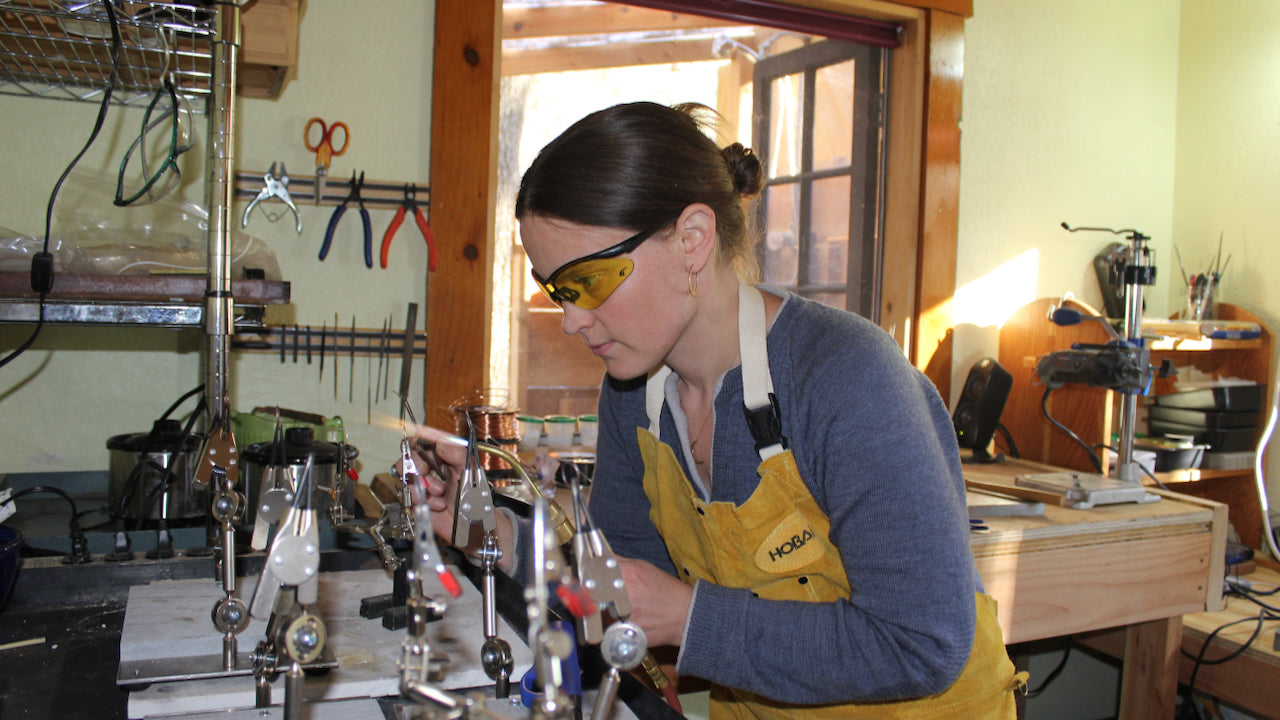 Welcome Inside My Studio' video featuring jeweler LothLorien Stewart wearing safety glasses and apron while working with metalsmithing tools in her Yosemite-area workshop. Text overlay describes the artisan jewelry-making process with 'Molten metal. Swinging hammers. Whirling polishers.