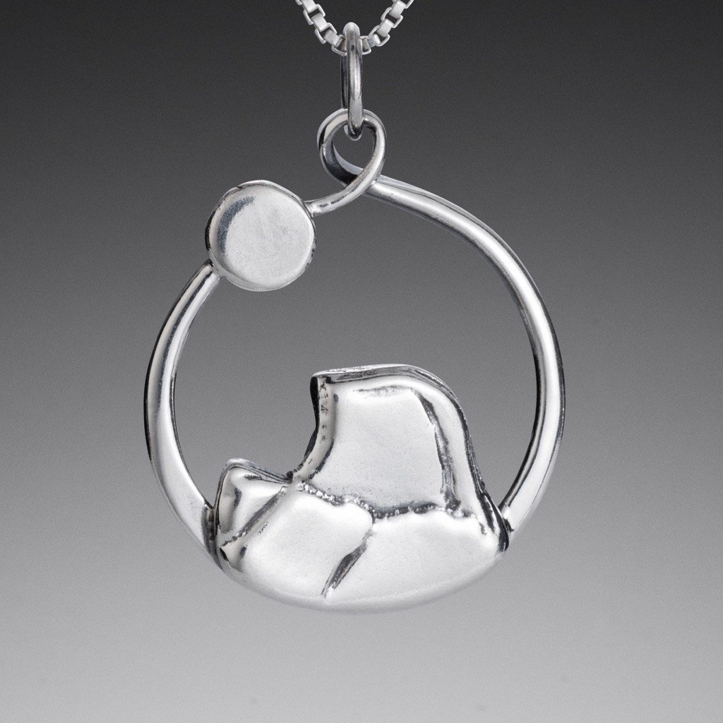 Sterling silver Half Dome pendant by Mostly Sweet Jewelry featuring a circular design with mountain silhouette inside. The pendant shows the distinctive profile of Yosemite's Half Dome with polished silver finish against gray background. Hangs from silver box chain with decorative bail attachment. Nature-inspired jewelry ideal for outdoor enthusiasts and national park lovers.