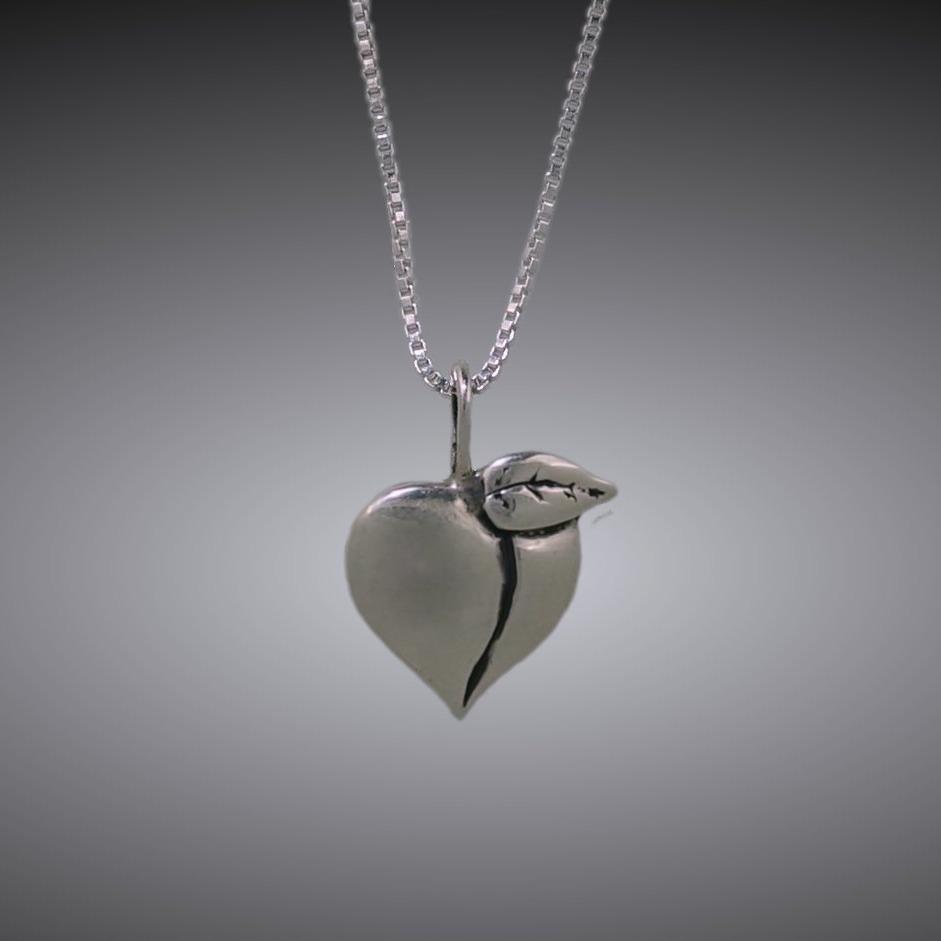 Sterling silver Oregon Country Fair Peach pendant by Mostly Sweet Jewelry. This handcrafted heart-shaped peach charm features a delicate leaf detail and polished finish. Shown hanging on a silver box chain against gray background. Commemorative jewelry piece celebrating Oregon's agricultural heritage and the iconic country fair.